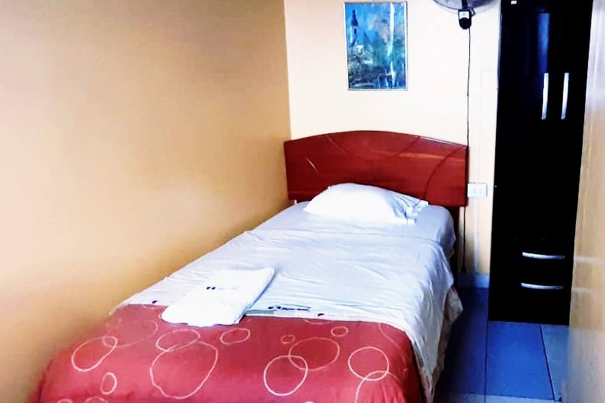 Una cama individual con cabecera roja y ropa de cama blanca, un cuadro pequeño en la pared y una puerta a la derecha.