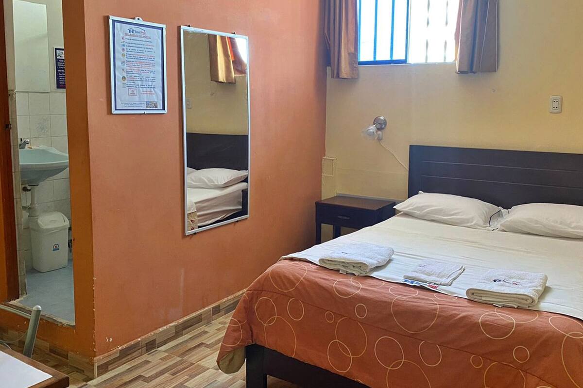 Una habitación de hotel con cama, mesita de noche, espejo, ventana con cortinas, lavamanos y un inodoro.
