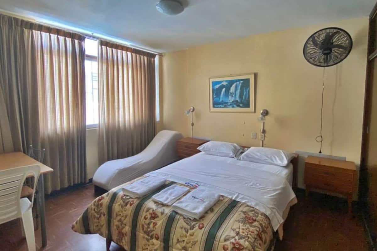 Una habitación de hotel con una cama, un escritorio, una silla, un ventilador y un cuadro en la pared.