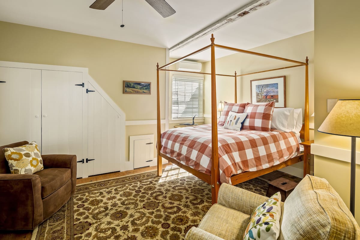 A bedroom with a four-poster bed, a brown sofa, a patterned rug, and a window with blinds.