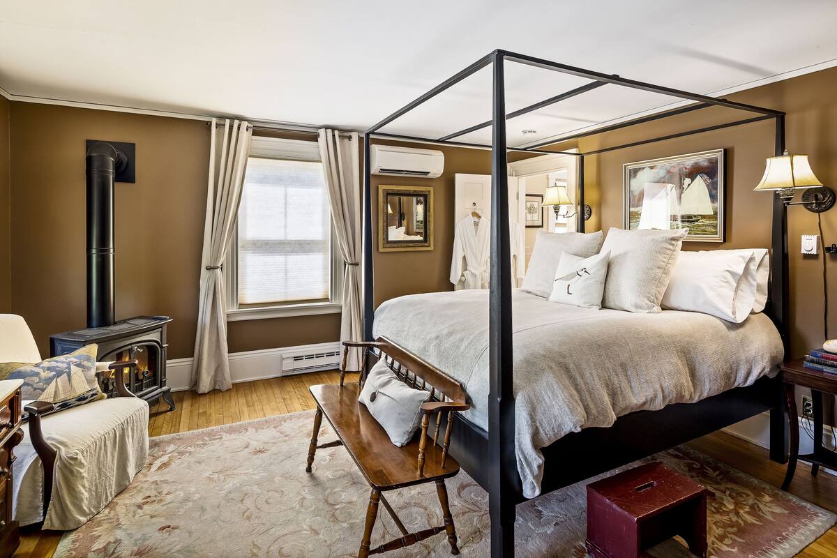 A four-poster bed with a white bedspread, a bench with a pillow, a red stool, a fireplace, and a large window with curtains.