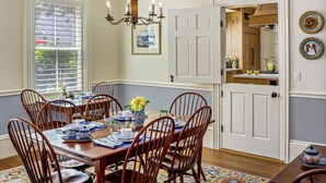 A dining room with a wooden table set for six, surrounded by wooden chairs. A chandelier hangs from the ceiling, and a window with blinds is visible. - Abigail's Inn (Camden)