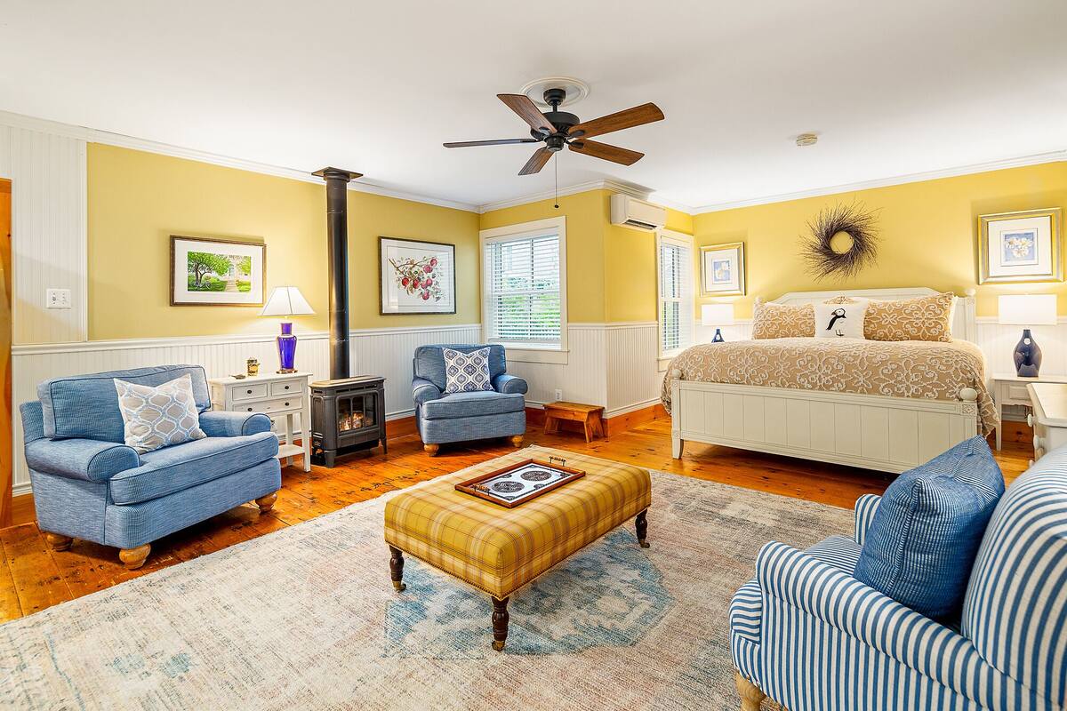 A room with a bed, two sofas, a small table, a floor lamp, a ceiling fan, and a fireplace.