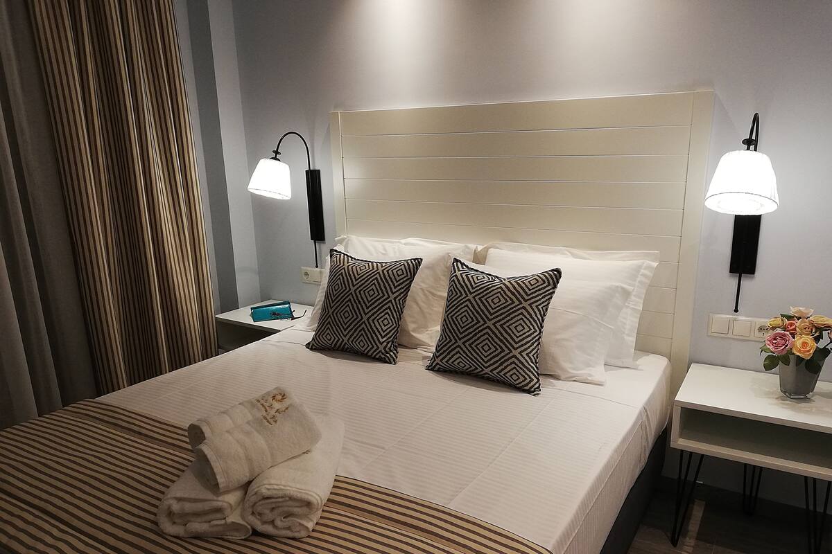 Een net opgemaakt bed met twee kussens, een nachtkastje met een lamp en een vaas met bloemen.