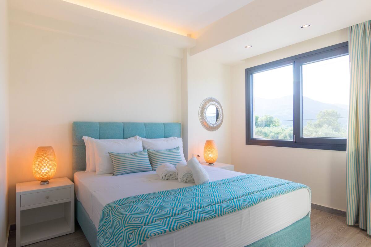 Een net opgemaakt bed met een geborreld sprei, twee bedlampjes en uitzicht op de omgeving door een groot raam.