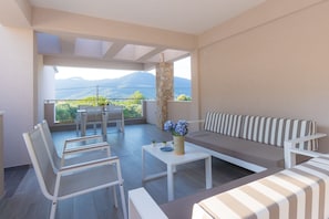 A modern living room with a sofa, chairs, and a coffee table. A balcony with a view of mountains is visible through the window. - Bella Vista Luxury Apartments (Thasos)