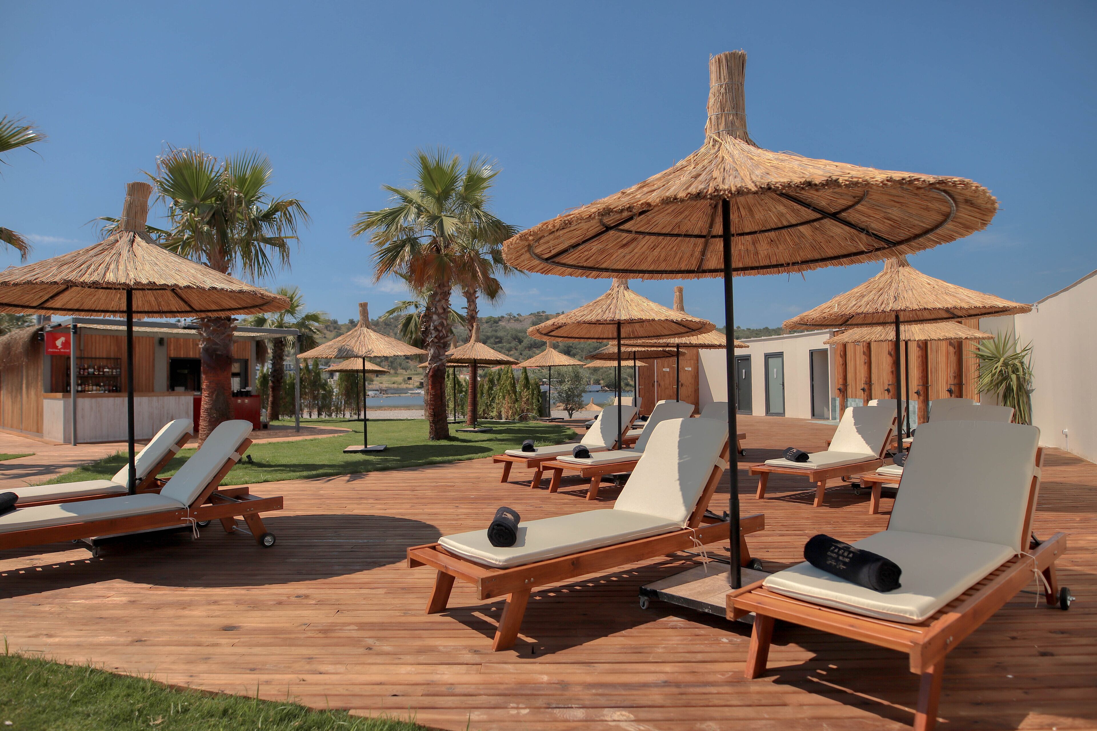 A wooden deck with sun loungers and thatched umbrellas, palm trees, and a building in the background.