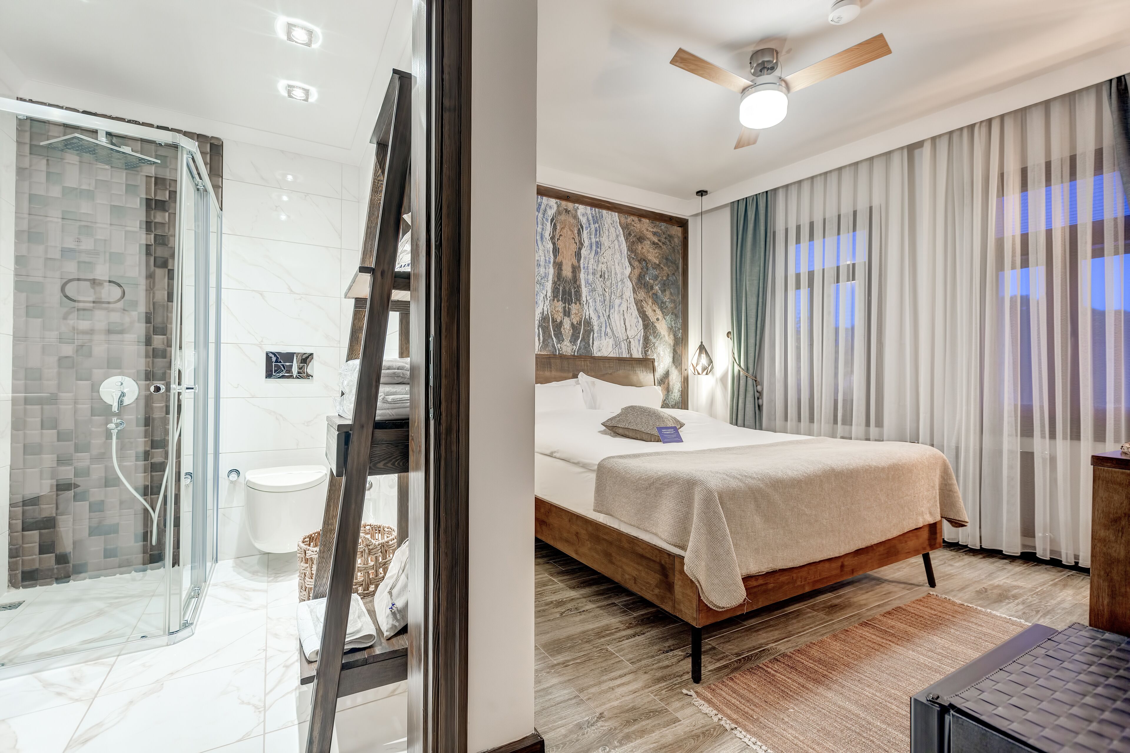 A modern hotel room with a glass-enclosed shower, a wooden bed, and a view of the city through sheer curtains.
