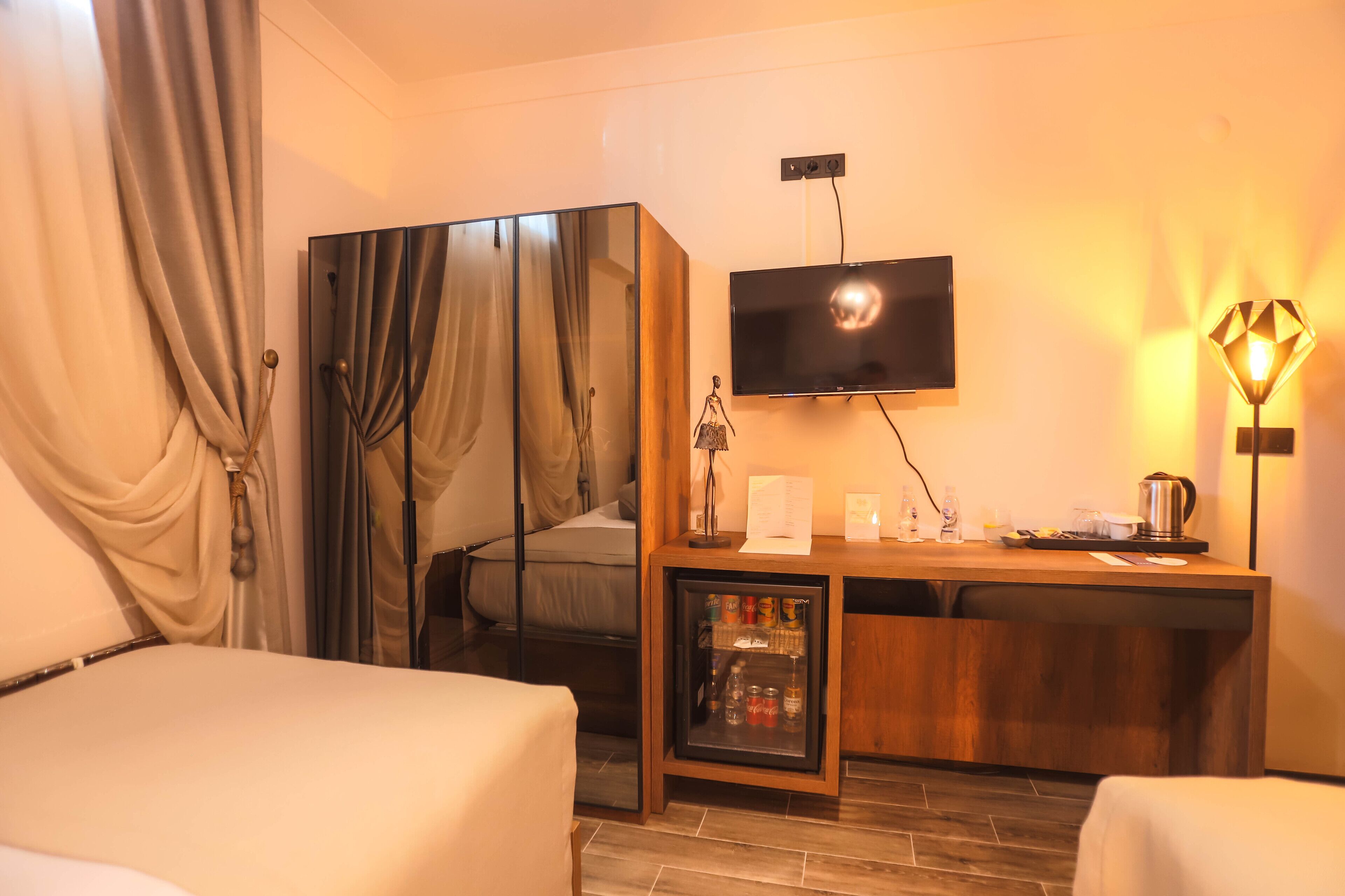 A hotel room with a bed, a desk with a television, a minibar, and a lamp.