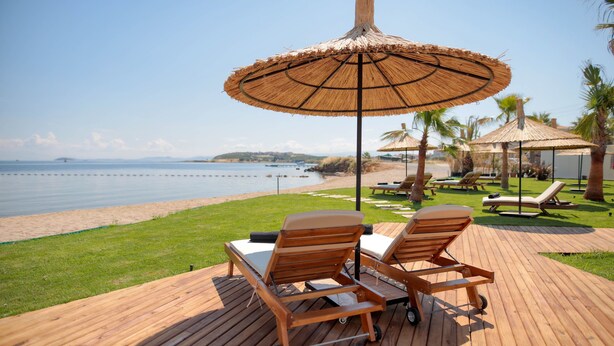 A wooden deck with two lounge chairs and a table, overlooking a beach with palm trees and a clear sky. - Parna Hotel (Ayvalik)