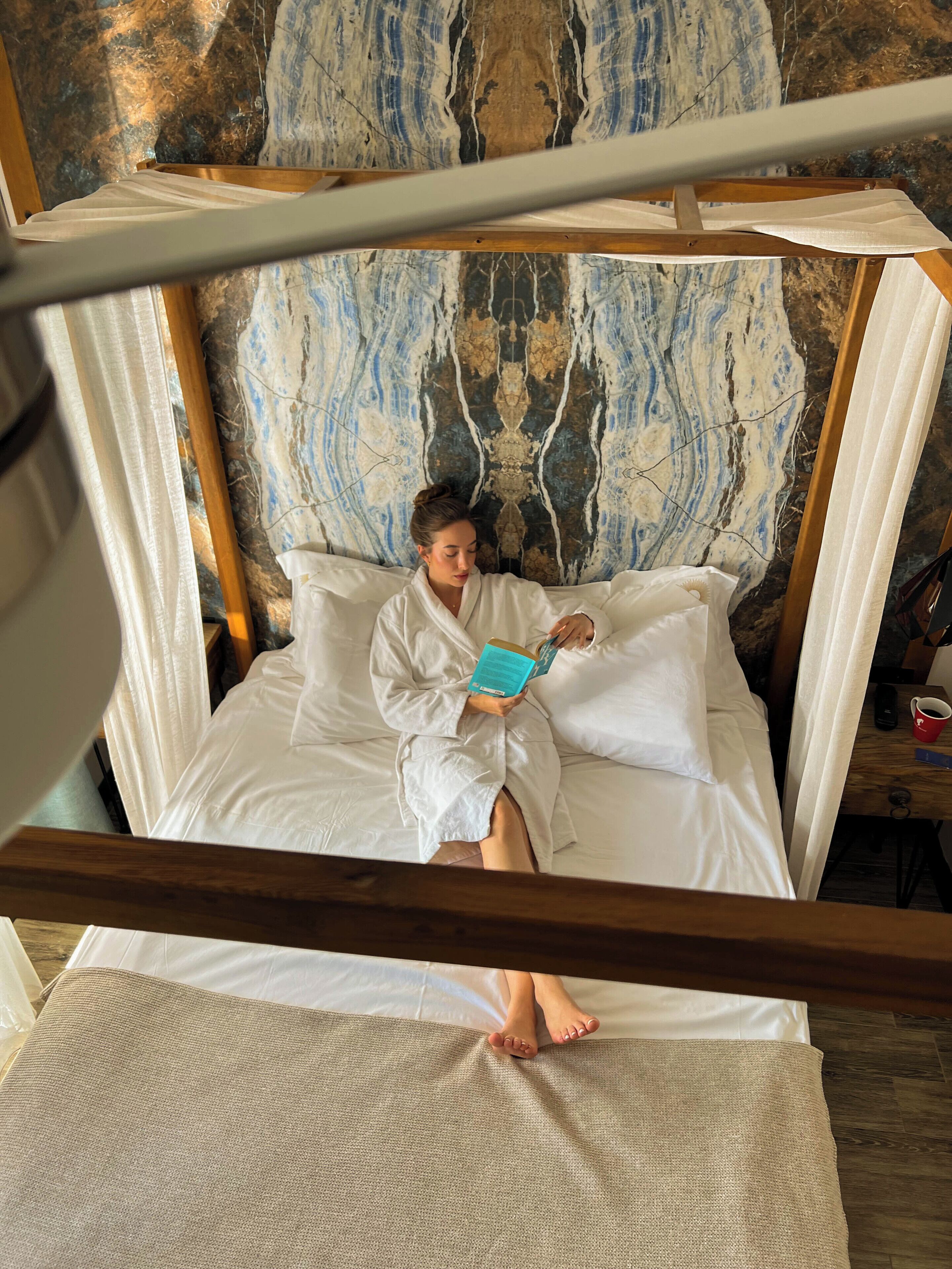 A person in a white bathrobe sitting on a bed with a marble-patterned headboard, reading a book.