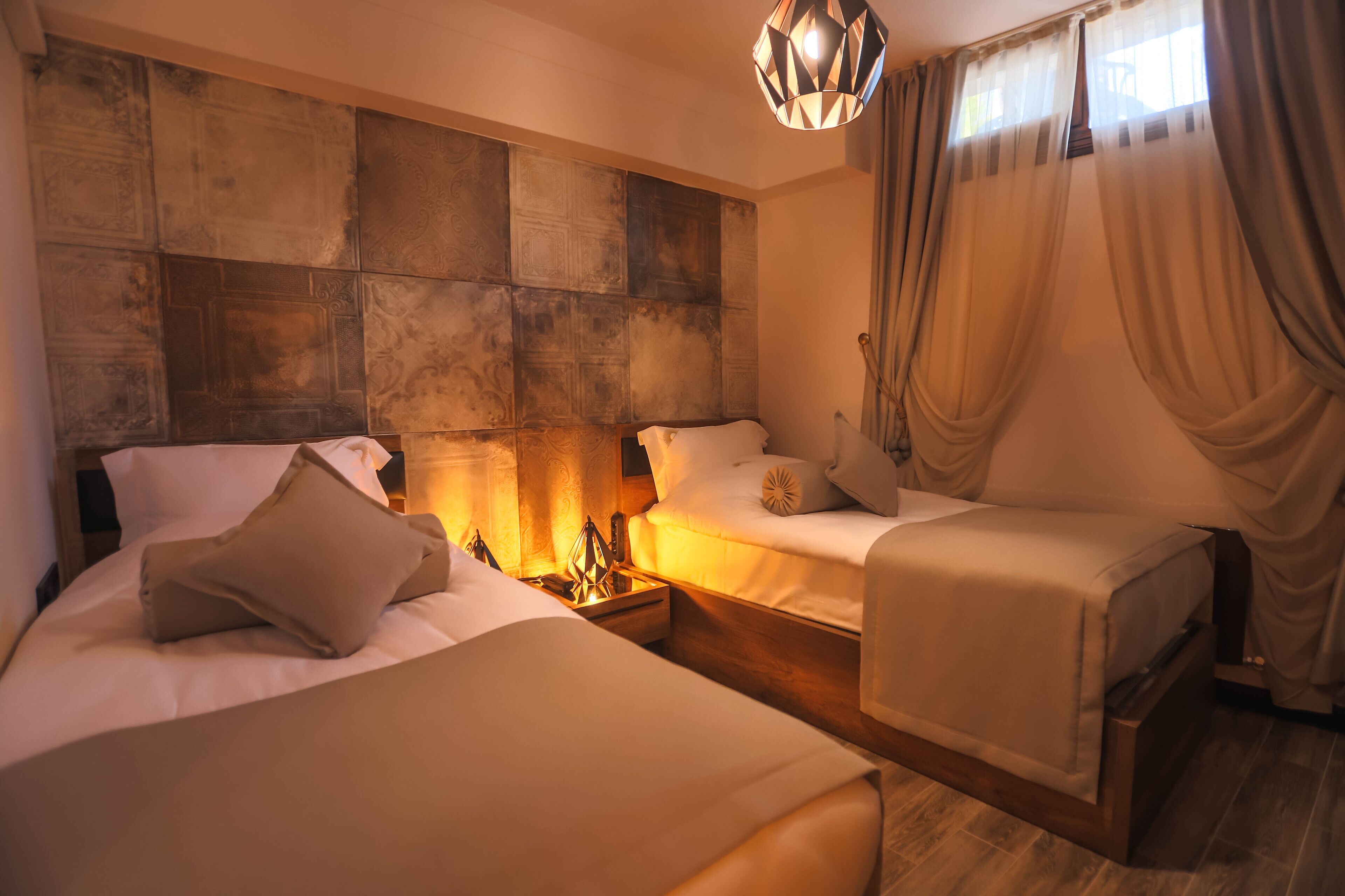 A hotel room with two beds, a decorative headboard, a nightstand with a lamp, and a window with curtains.