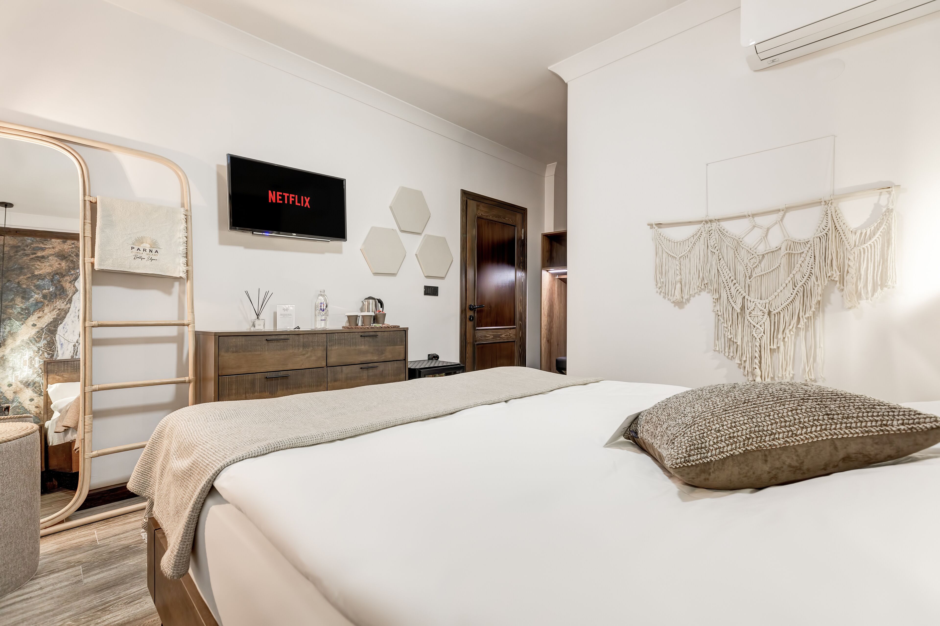 A modern bedroom with a bed, a TV mounted on the wall, a wooden dresser, and a macrame wall hanging.
