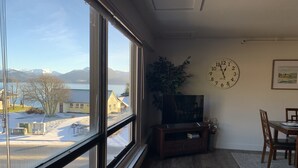 A room with a large window offering a view of a snowy landscape and mountains. There is a wooden entertainment unit with a television, a dining table with chairs, and a wall clock. - Downtown Apt. 5 w/ Ocean & Mountain Views. (Sitka)