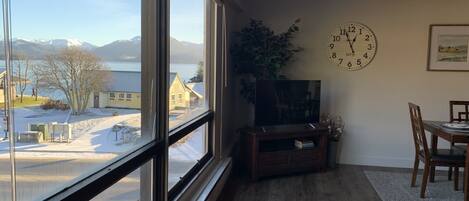 Een kamer met een groot raam dat uitzicht geeft op een besneeuwd landschap en bergen. Er is een houten dressoir met een televisie, een eettafel met stoelen en een wandklok.