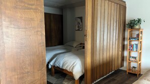A bedroom with a wooden wardrobe, a bed, a bedside table with books, and a potted plant. - Downtown Apt. 5 w/ Ocean & Mountain Views. (Sitka)