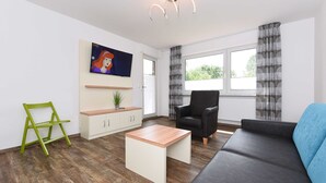 Una sala de estar moderna con un sofá, una mesa de centro, una silla, un televisor montado en la pared y una ventana con cortinas.
