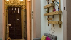 A lobby with a wooden reception desk, a grey sofa with a yellow pillow, a wooden shelf with vases, and a chandelier. - Maria Bonita - Hostel (Guadalajara)