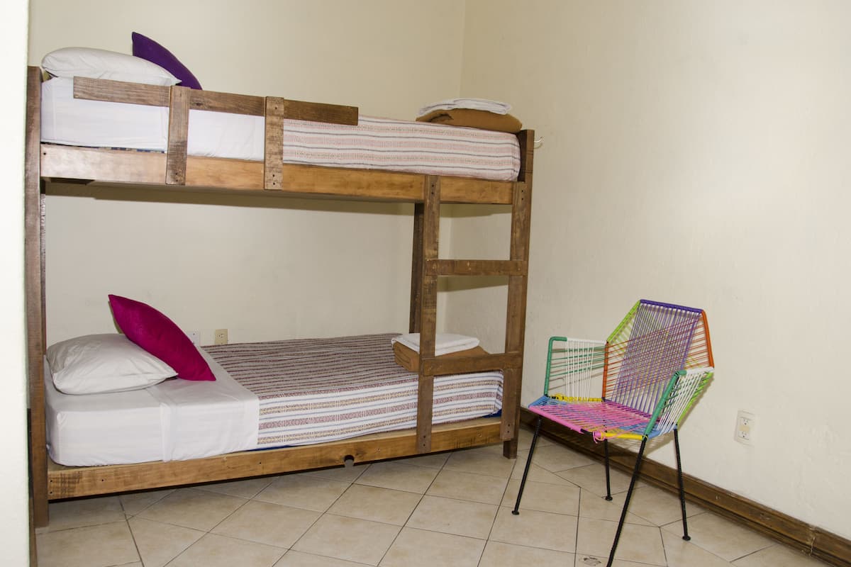 Una cama litera con estructura de madera y ropa de cama blanca, una silla colorida y una pared sencilla.