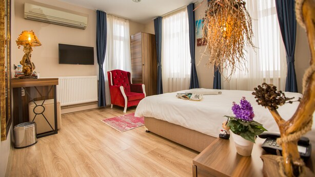 Ein Hotelzimmer mit einem Bett, einem roten Sessel, einem Holztischchen, einer Lampe, einem Fernseher und einem Fenster mit Vorhängen.