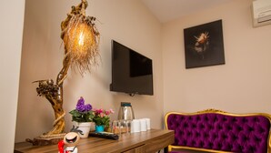 Ein Zimmer mit einem Holztisch, einem an der Wand befestigten Fernseher, einem violetten Polsterstuhl, einer Zierpflanze und einer außergewöhnlichen Pendelleuchte.