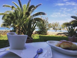 Una mesa con una taza blanca, una cuchara y un plato con cruasanes, ubicada al aire libre con palmeras y vistas al mar.