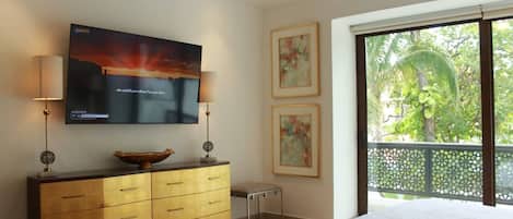 A bedroom with a bed, a television mounted on the wall, a wooden dresser, and a window with a view of greenery.