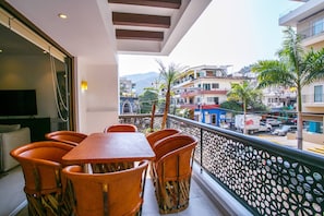 Een balkon met een tafel en stoelen, uitkijkend op een straat met gebouwen en palmbomen.