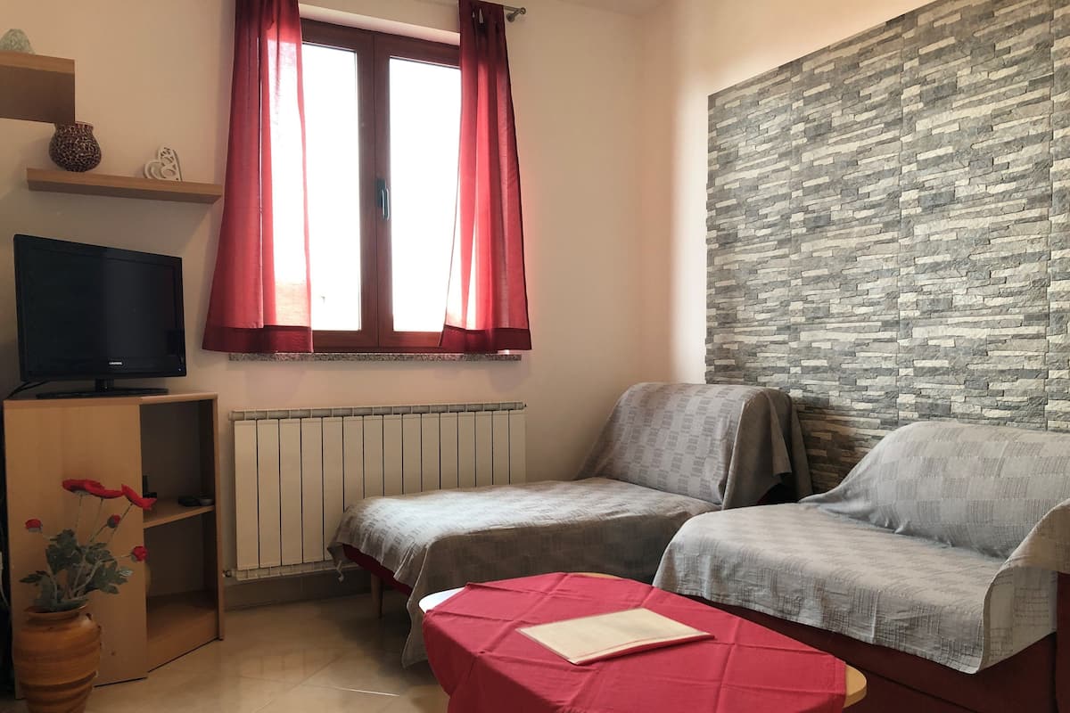 Ein kleiner, gemütlicher Raum mit einer Couch, einem Fernseher, einem Fenster mit roten Vorhängen, einer Steinwand und einem Holzbodenregal mit Dekoration.