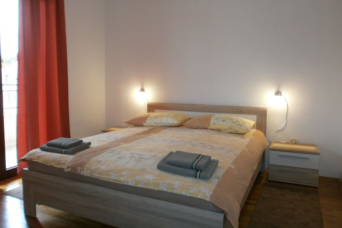 Ein ordentlich bezogenes Bett mit floralem Überwurf, zwei Nachttischlampen und ein Nachttisch aus Holz mit einer Schublade.