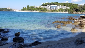 A coastal view with clear blue waters, rocky shoreline, and a building with multiple balconies overlooking the sea.