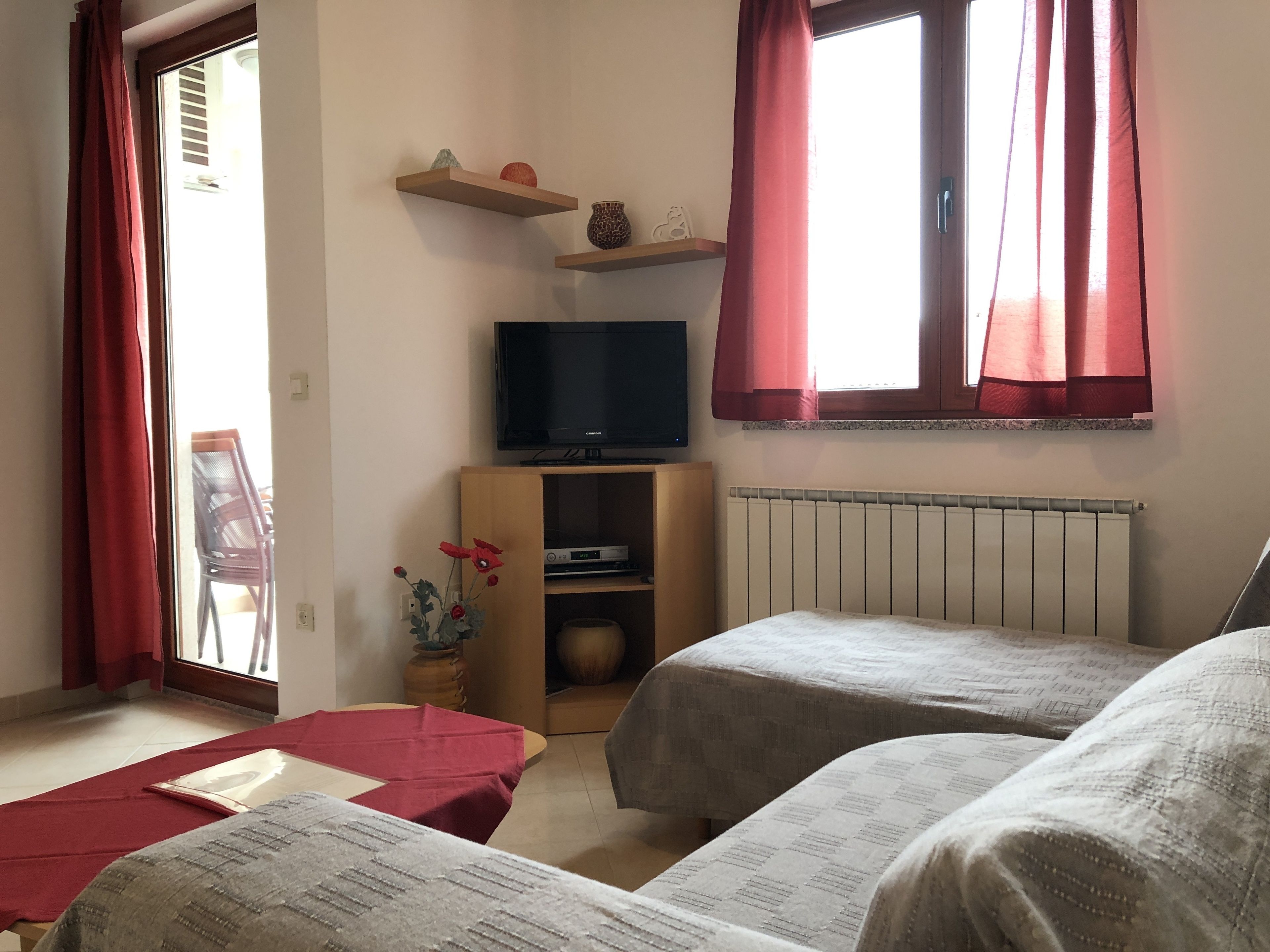 A hotel room with two beds, a television on a wooden shelf, a radiator, and a window with red curtains.