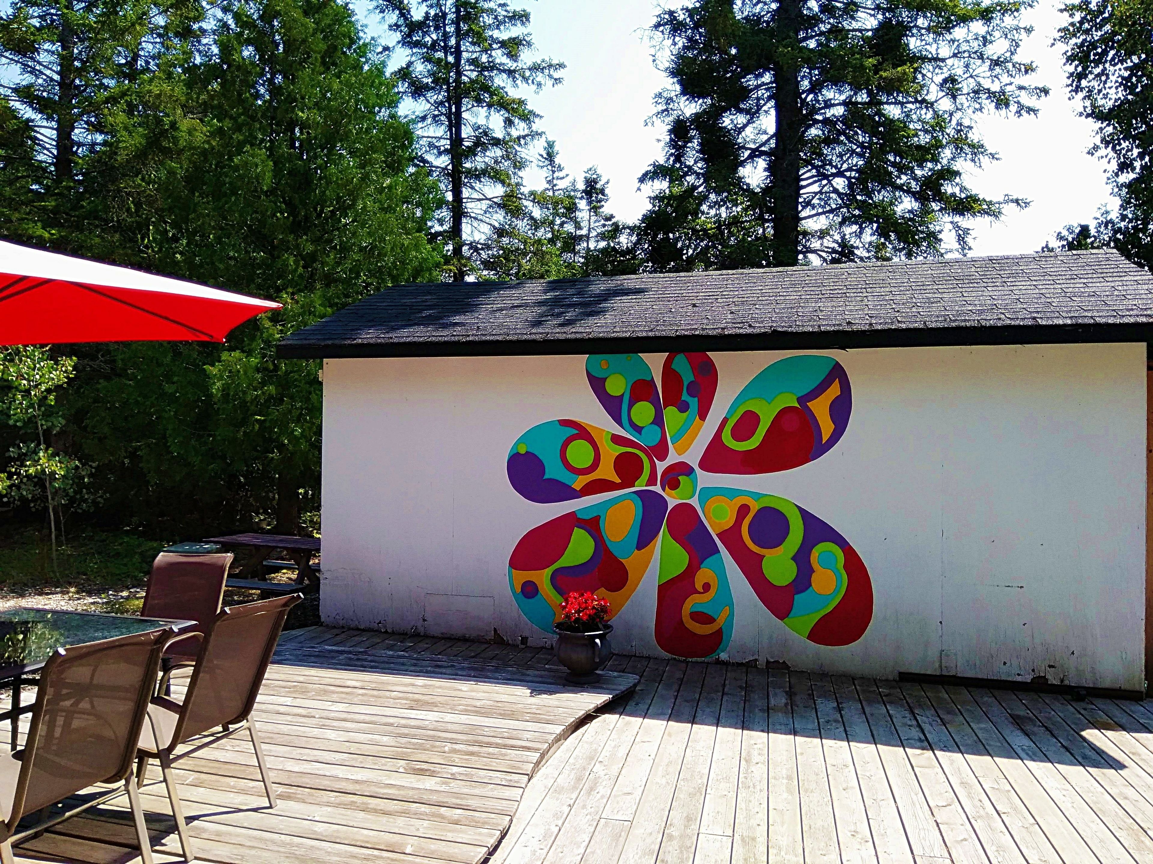 A wooden deck with a colorful flower mural on a building wall, outdoor seating, and a red umbrella.