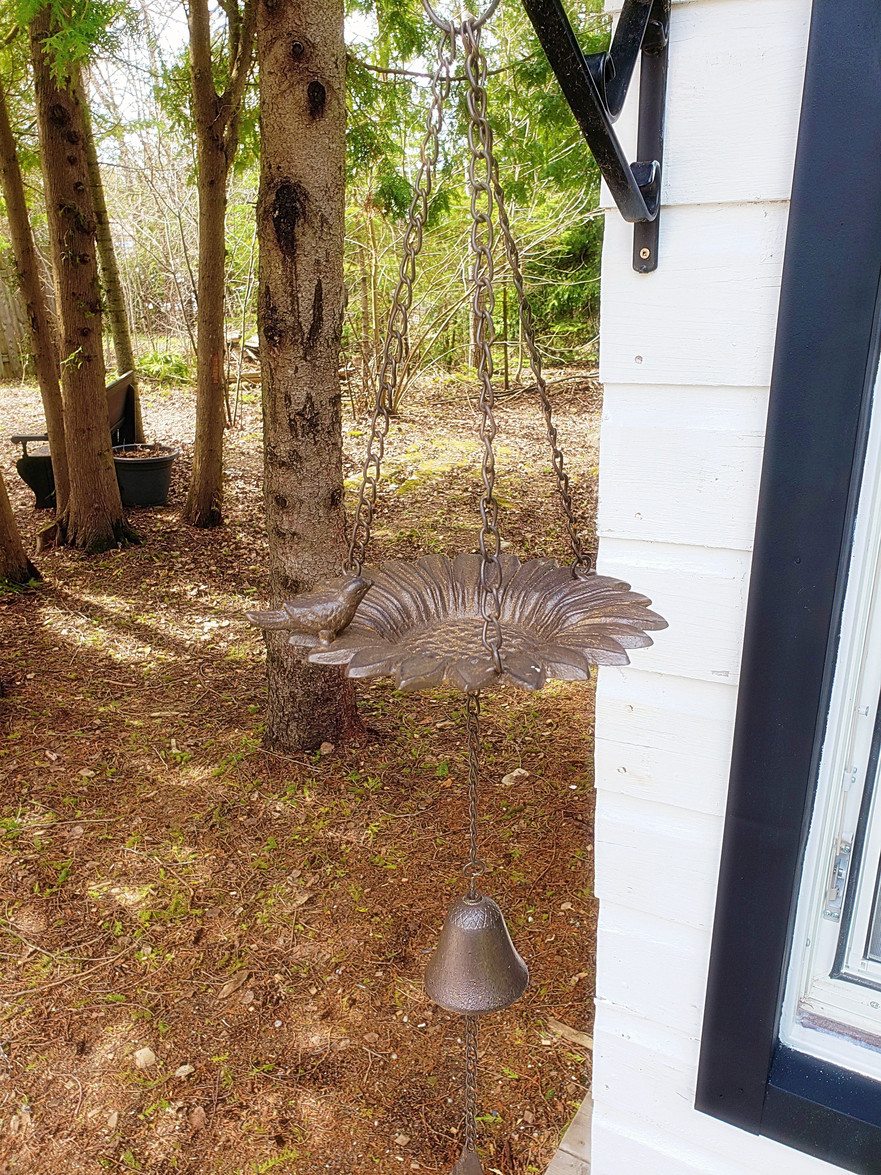 A rustic outdoor area with a metal birdbath and a small table, surrounded by trees and a white building.