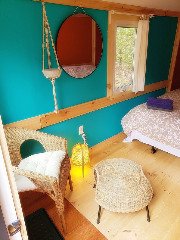 Ein Zimmer mit einer türkisfarbenen Wand, einem runden Spiegel, einem Bett mit gemustertem Überwurf, einem Korbstuhl, einem kleinen runden Tisch und einer hängenden Makramee-Lampe.