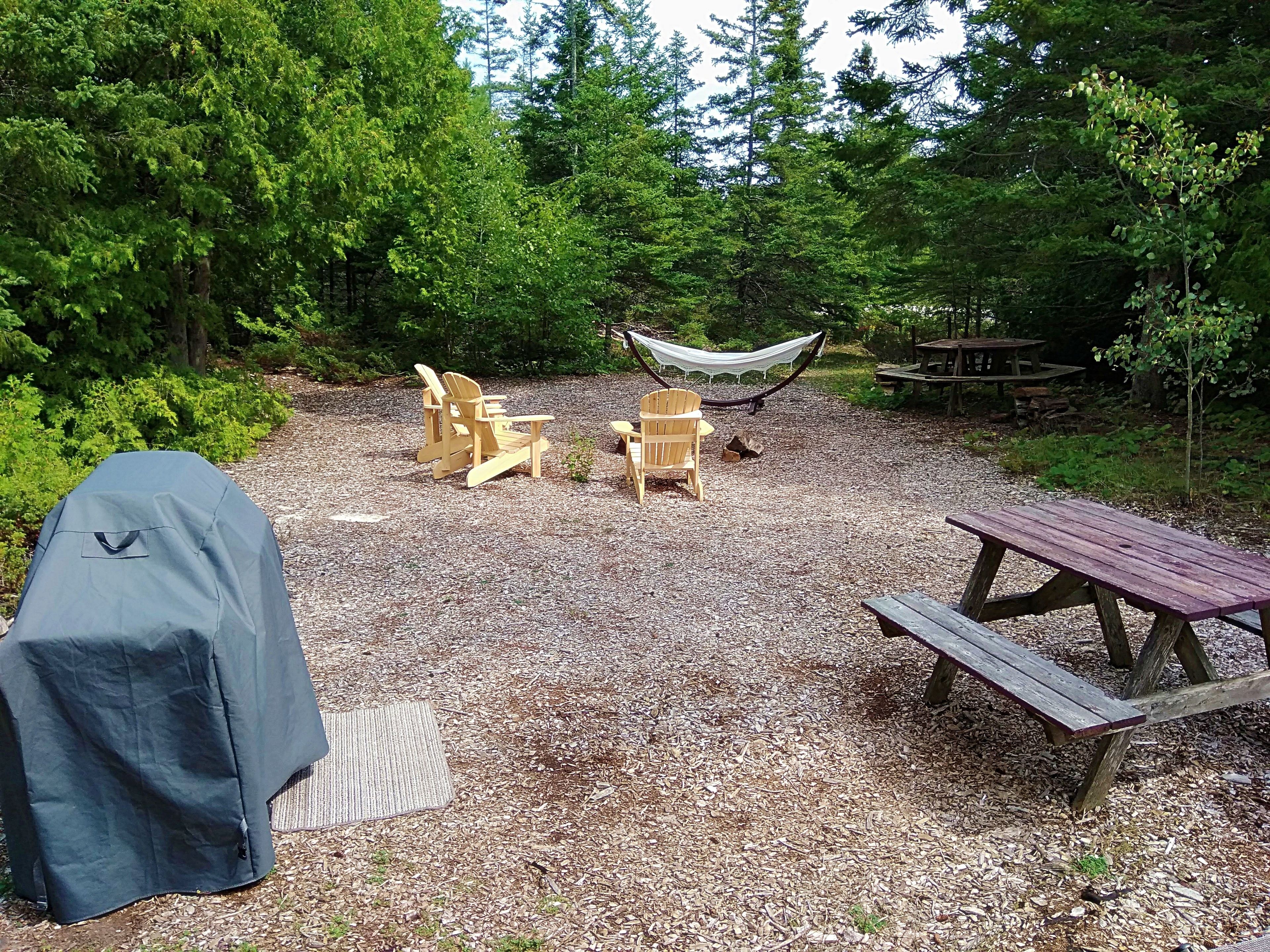A covered picnic area with a grill, two Adirondack chairs, a hammock, and a wooden picnic table.