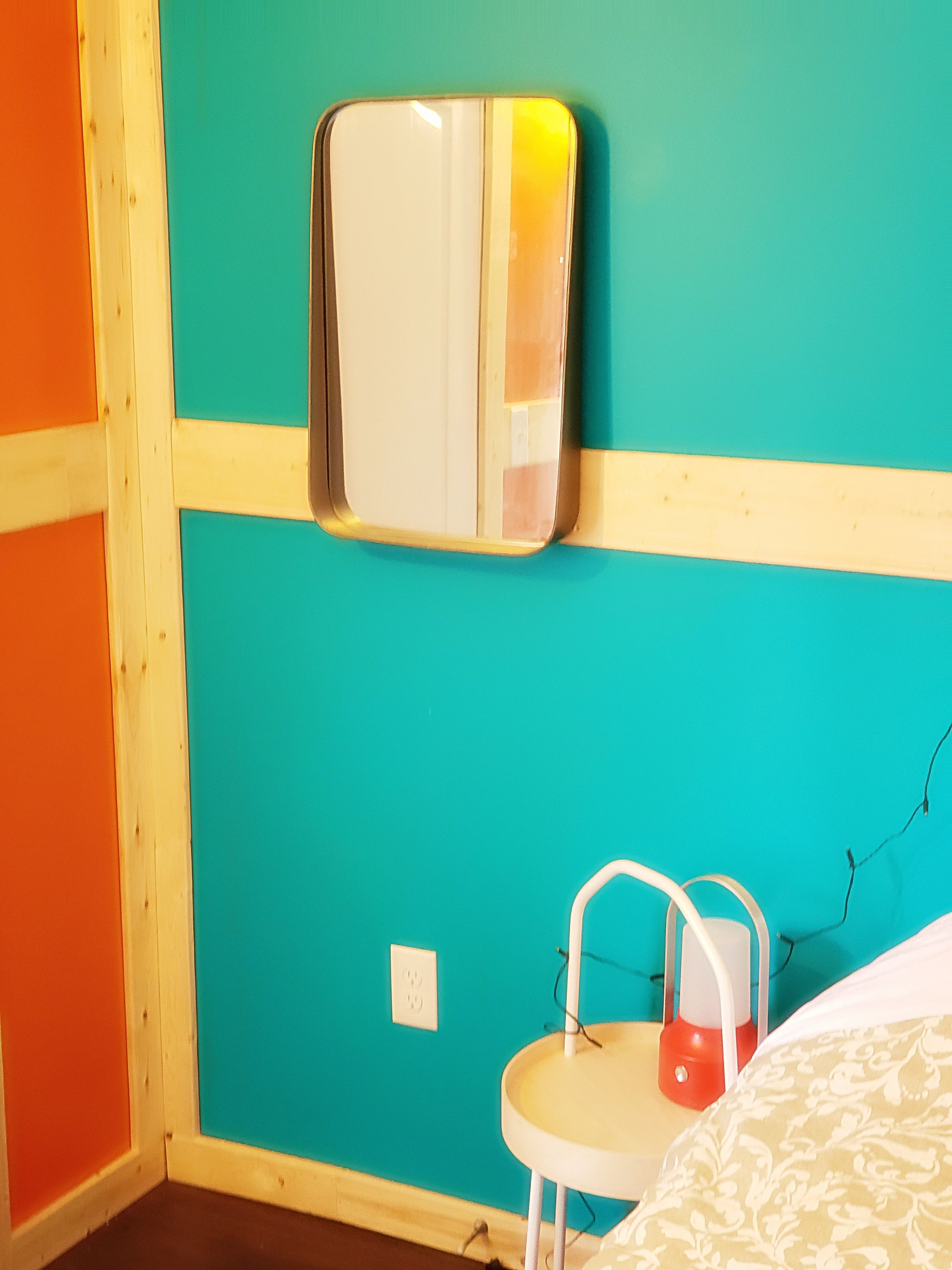 A room with teal walls, a wooden floor, a small white table with a red lamp, and a mirror on the wall.