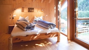 Ein Schlafzimmer in einer Holzhütte mit einem Bett, Kissen, einer Nachttischlampe und einem Fenster mit Blick auf eine Holzterrasse und Bäume.