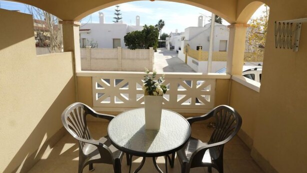 Un balcon avec une table ronde et deux chaises, une balustrade blanche et une vue sur une rue résidentielle.