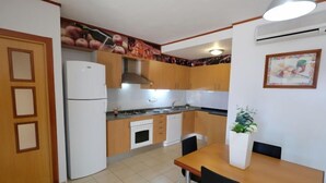 Een kleine keuken met houten kastjes, een witte koelkast, een fornuis en een spoelbak. Er staat een eettafel met stoelen en een potplant.