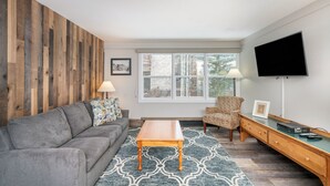 Un salon moderne avec un canapé gris, une table basse en bois, un tapis à motifs, un fauteuil, un téléviseur et une fenêtre donnant sur des arbres.