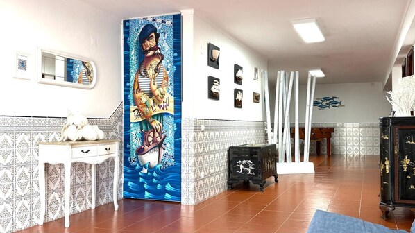 A room with a tiled floor, a white wall with a mosaic, a black cabinet, and a painting of a man on a boat.