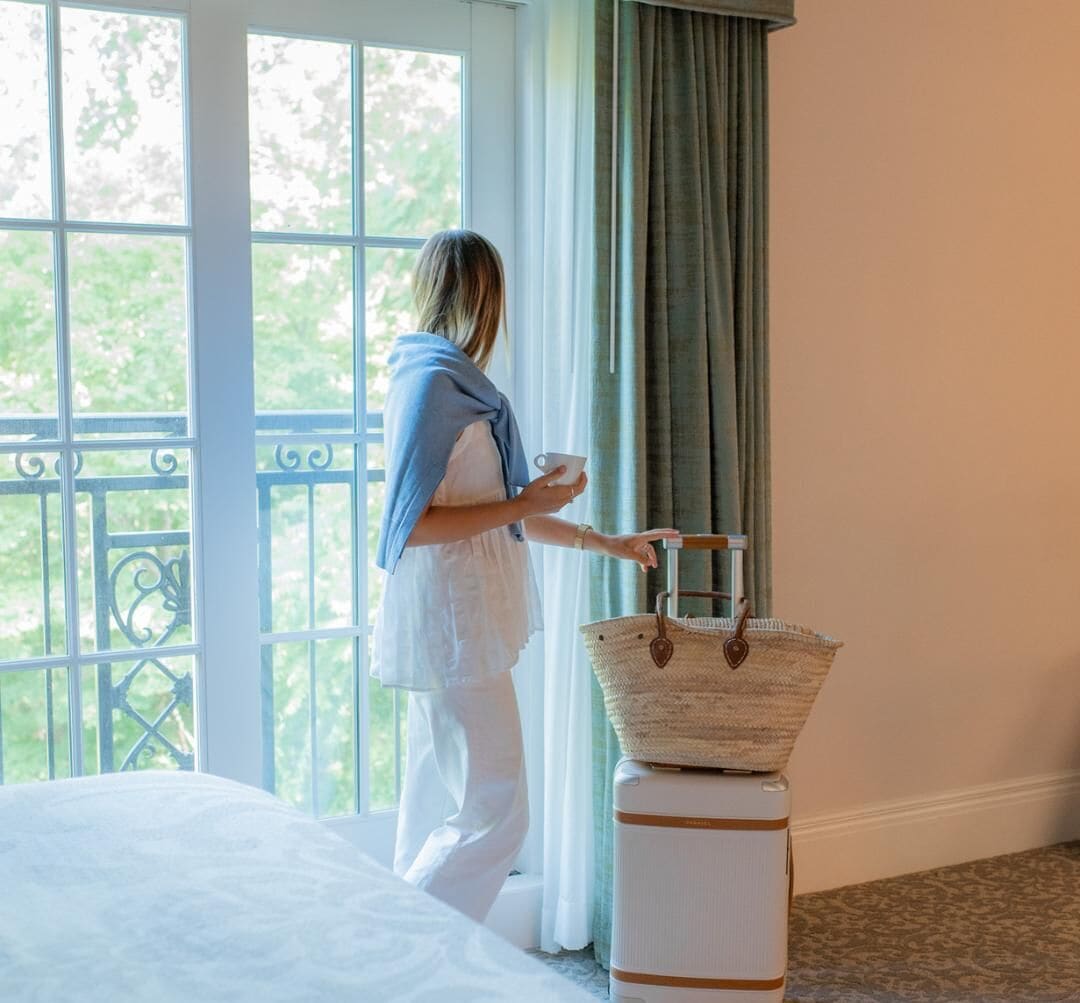 Une chambre avec un lit, une fenêtre avec des rideaux et une valise sur laquelle est posé un sac en tissu.
