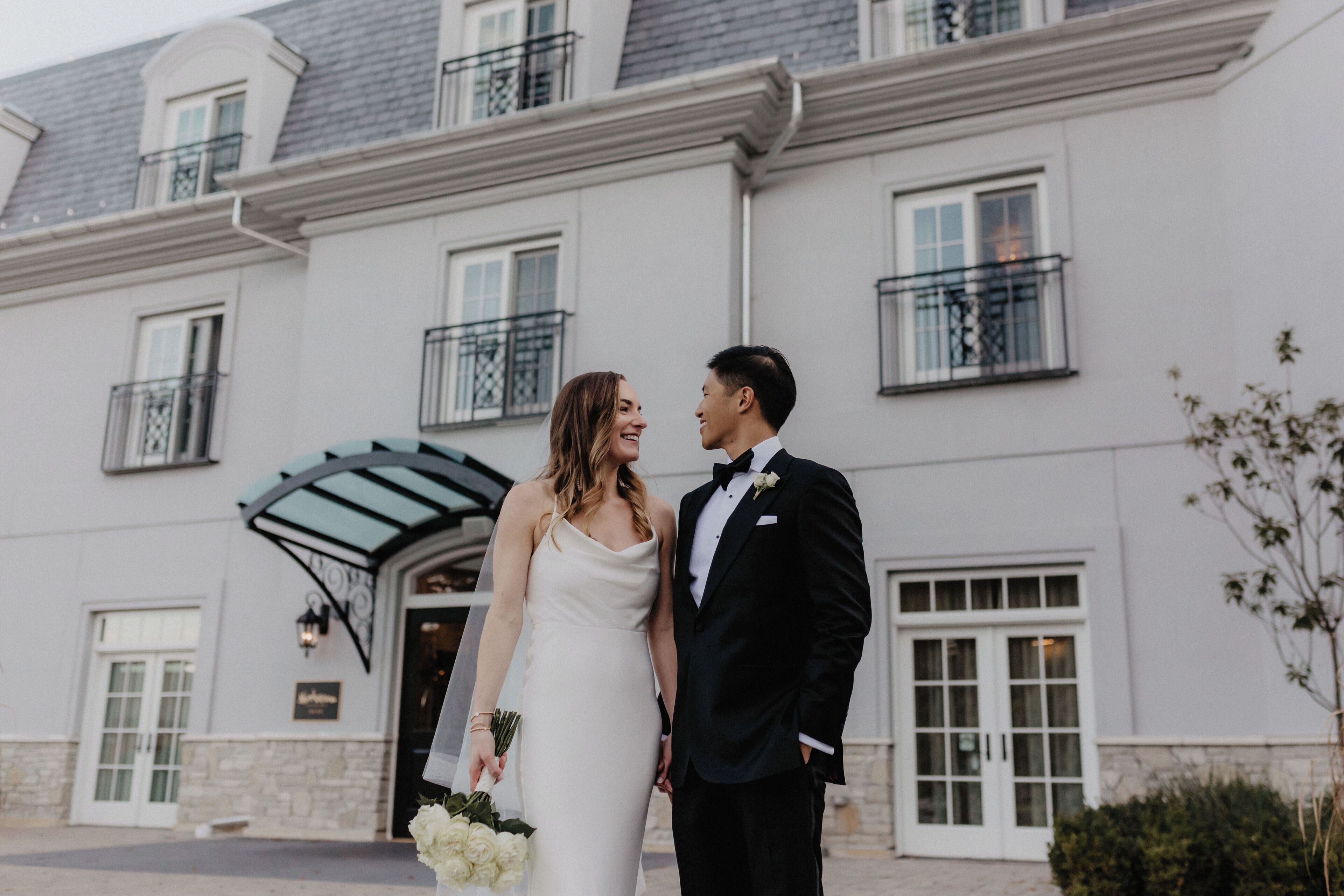 A couple in wedding attire standing in front of a building with a stone facade, large windows, and a balcony.