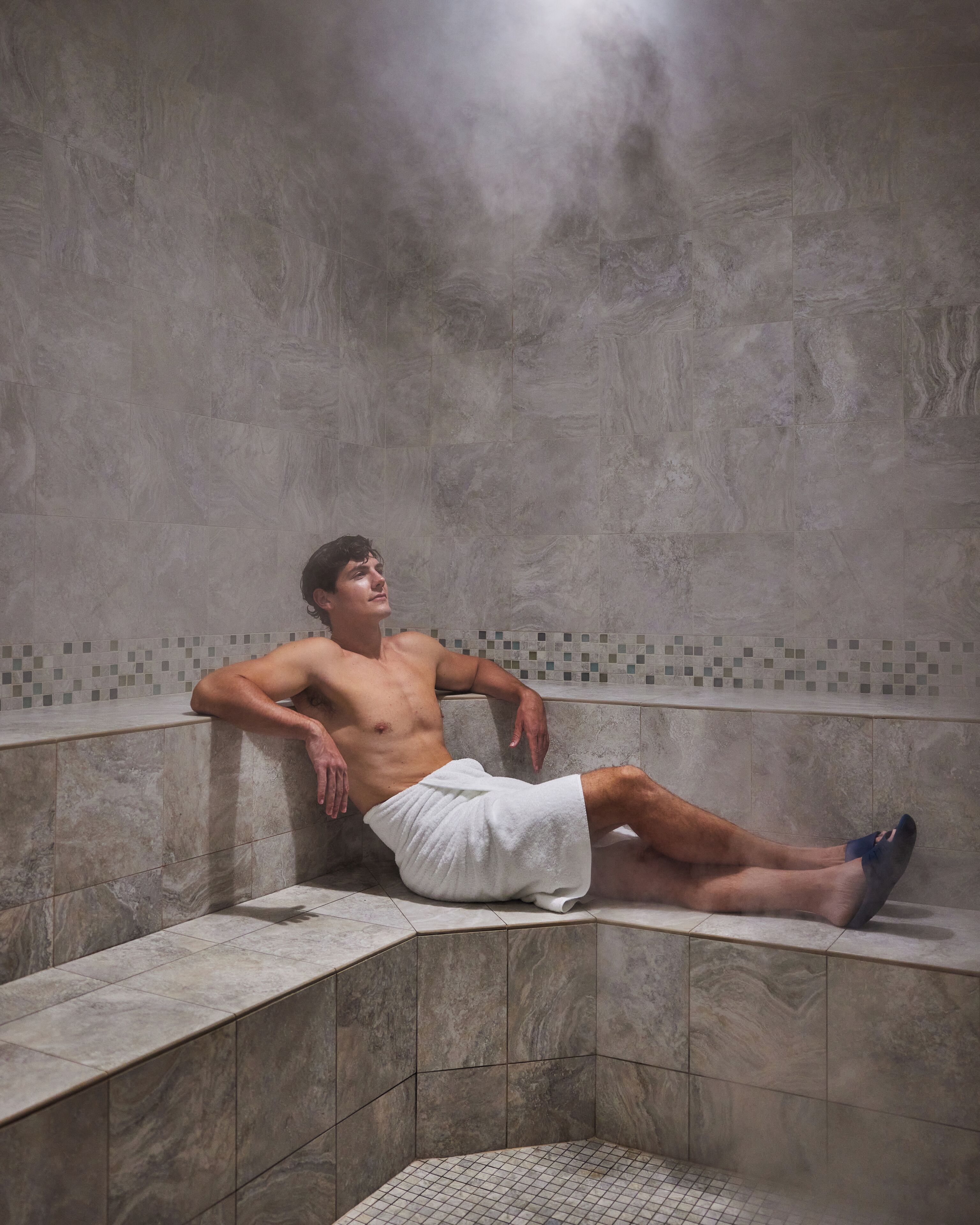 A person sitting in a steam room with tiled walls and a tiled floor.