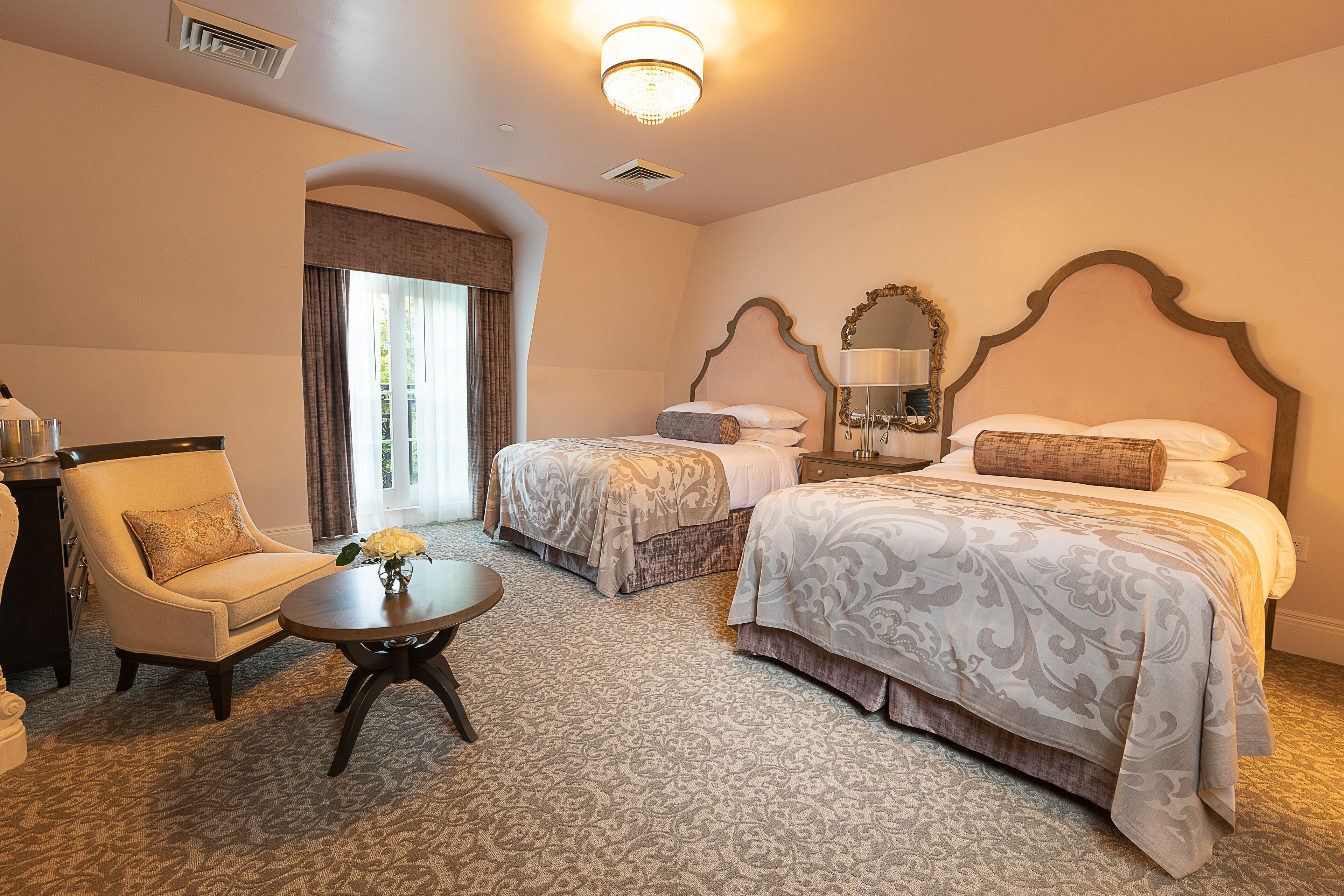 A hotel room with two beds, a chair, a small table with a vase, and a large window with curtains.