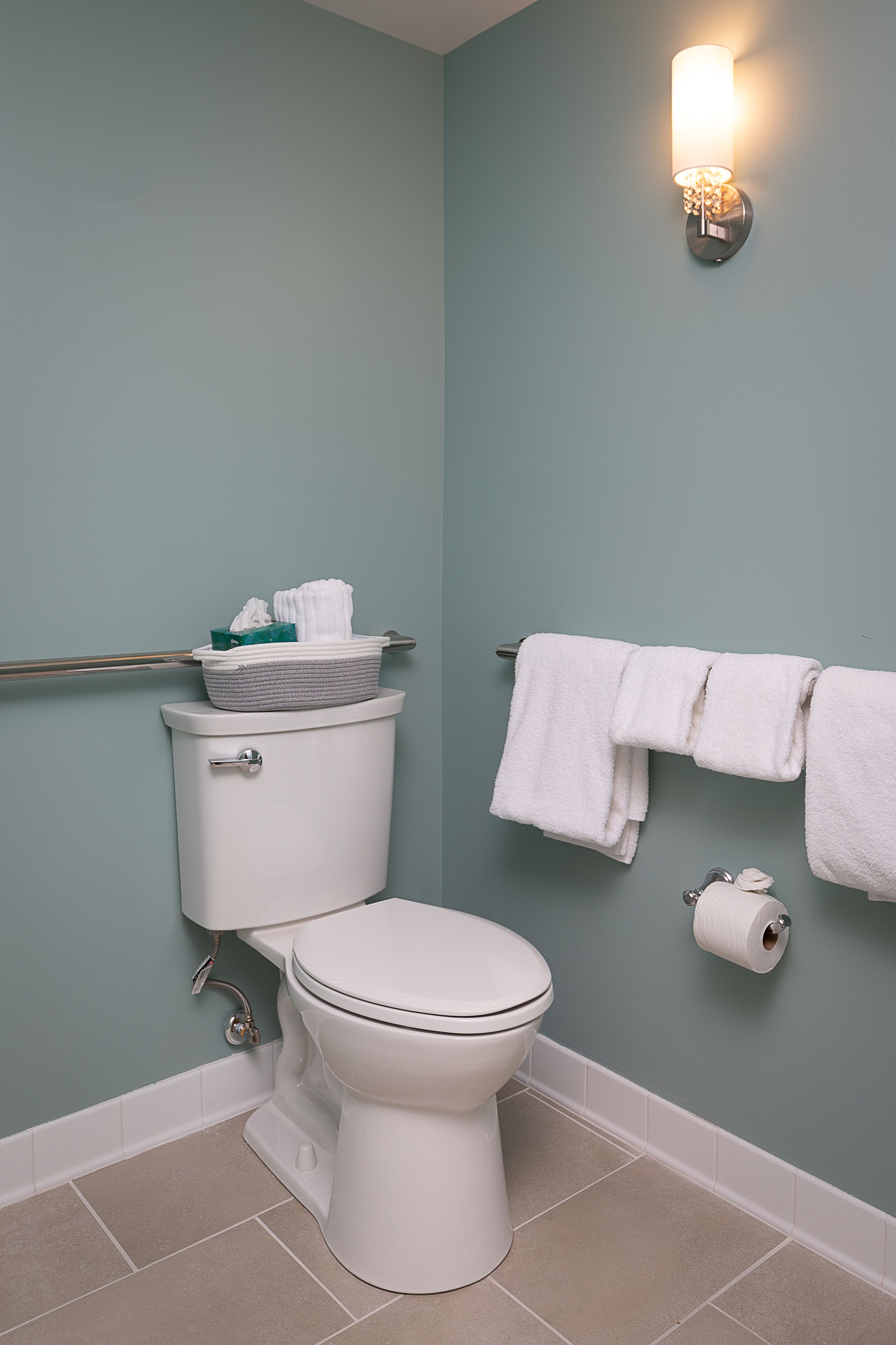 A bathroom with a toilet, a towel rack with towels, a toilet paper holder, and a wall-mounted light fixture.