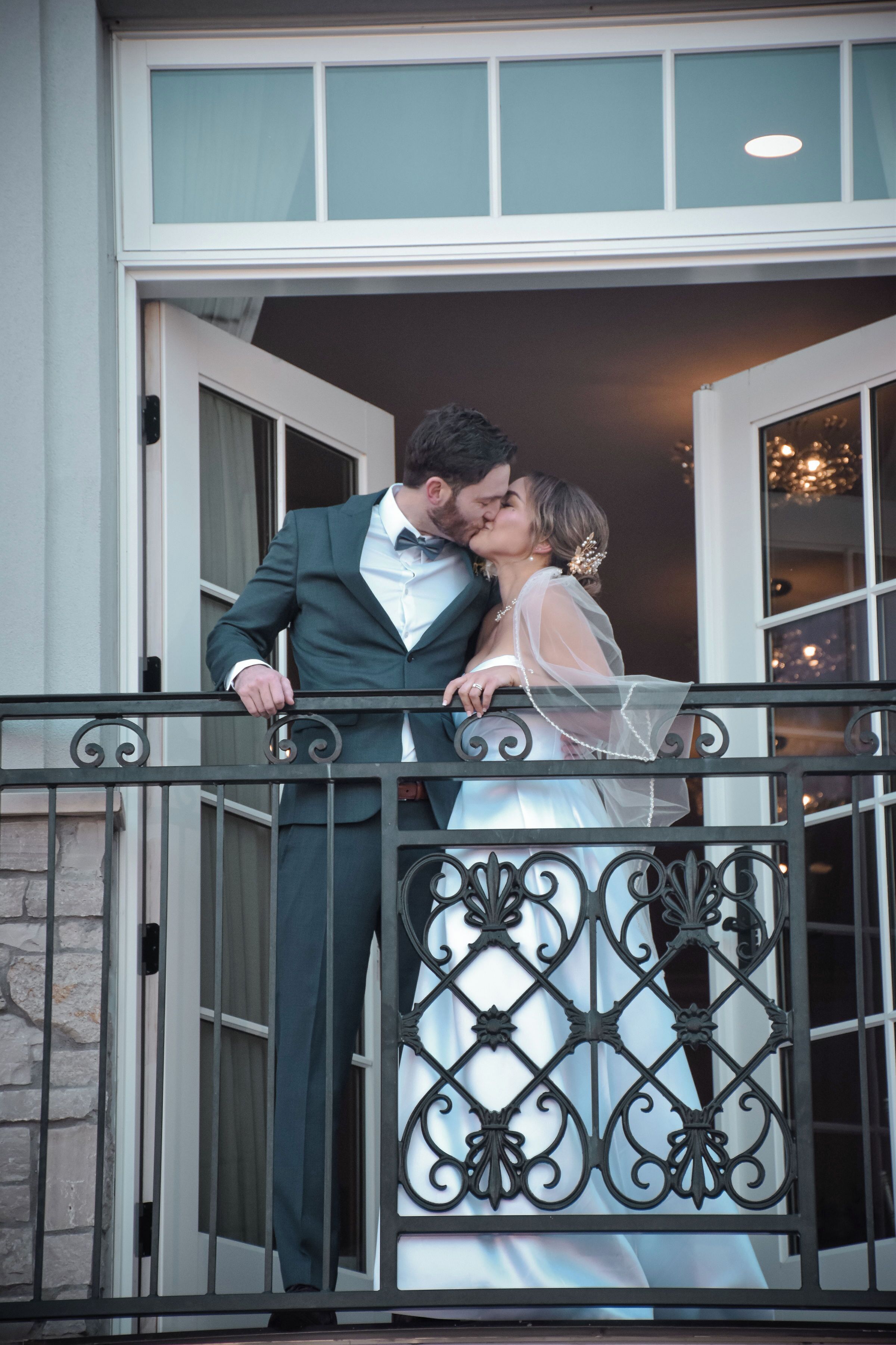 A couple on a balcony with a wrought iron railing, a stone wall, and a building entrance with glass doors.