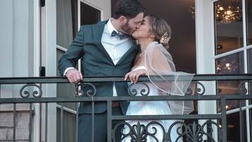 A couple on a balcony with a wrought iron railing, a stone wall, and a building entrance with glass doors.