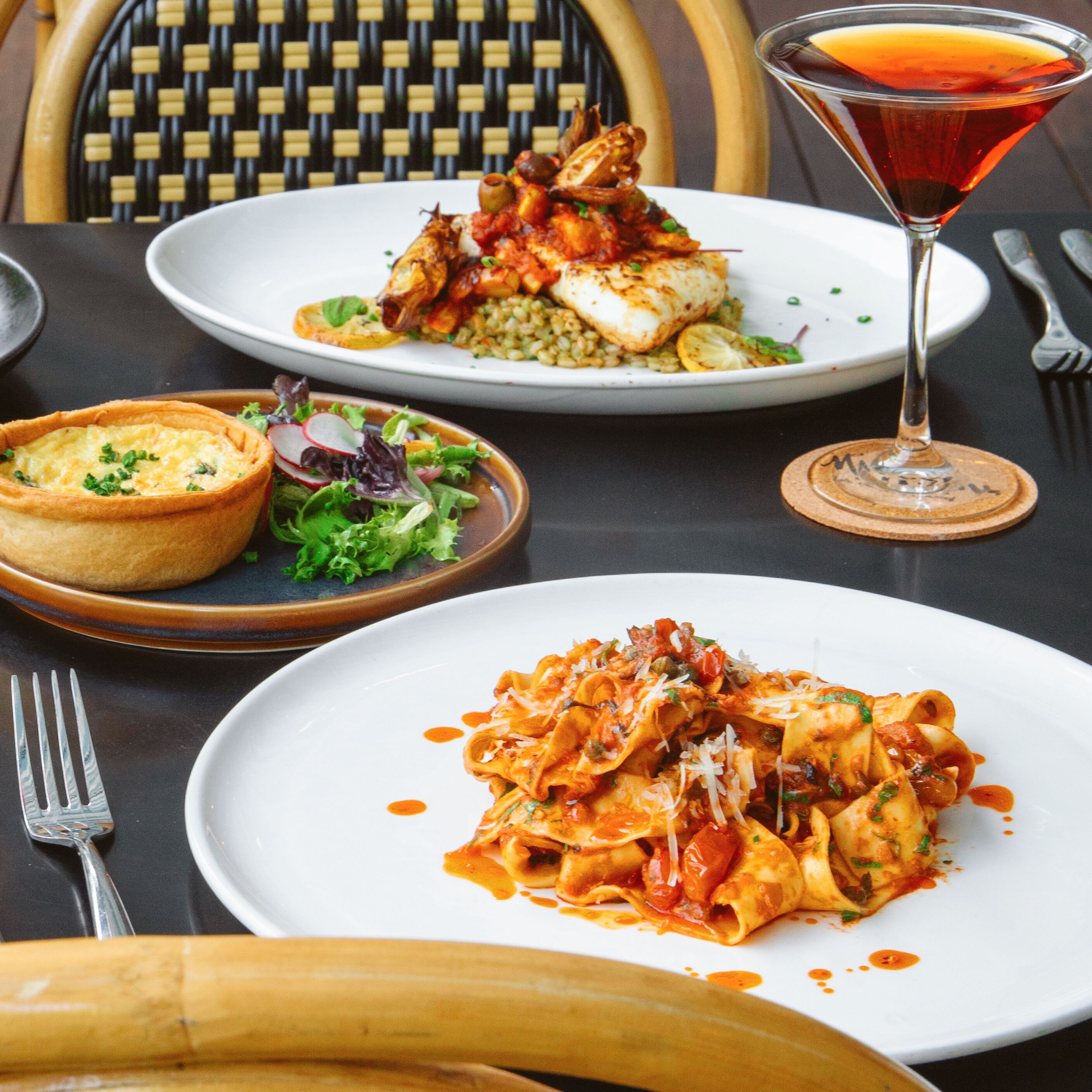 A dining table with plates of pasta and a seafood dish, a cocktail glass, and a bowl of salad.